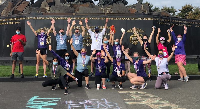 An enthusiastic group of runners in 261 Fearless shirts pose with raised arms and masks in front of a black wall with golden trophies 