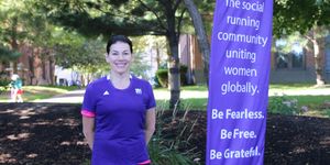 A woman in a purple 261 Fearless running shirt stands smiling next to a banner of the 261 Fearless running group