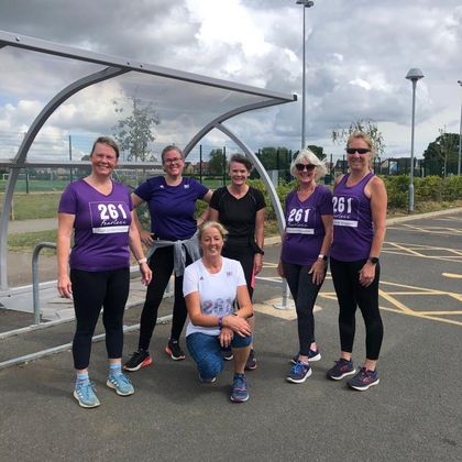Group photo of six women from the 261 Fearless running group on an empty sports field, posing in front of a bus stop, all wearing purple and blue running shirts