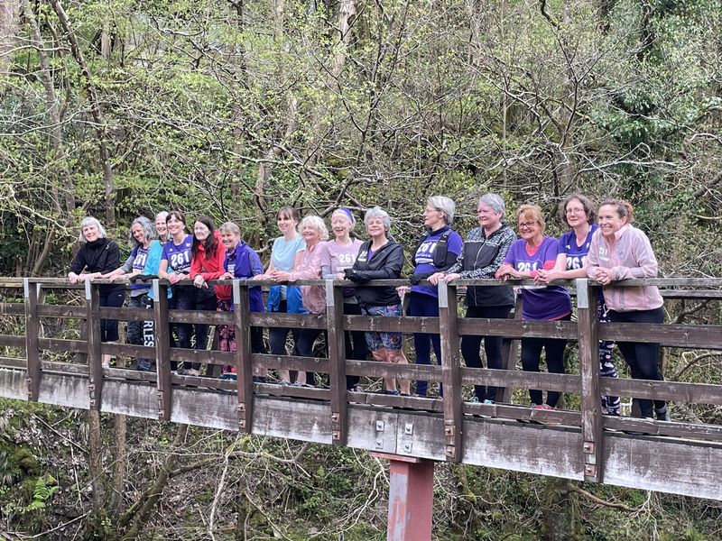 Group photo of numerous women of the 261 Fearless running group posing on a wooden bridge over a stream, surrounded by dense greenery, in a variety of running outfits
