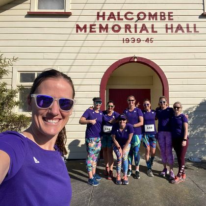 A group of cheerful runners in purple 261 Fearless T-shirts take a selfie in front of the historic Halcombe Memorial Hall, sun-drenched and full of energy 