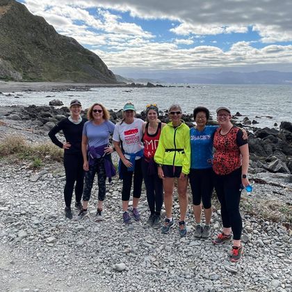 A group of eight 261 Fearless runners pose on the rocky shore of a beach, with the sea and steep hills in the background