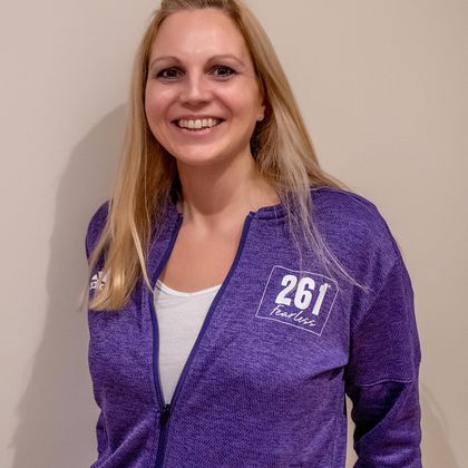 Portrait of a smiling woman in a purple 261 Fearless jacket proudly displaying her club logo against a neutral background
