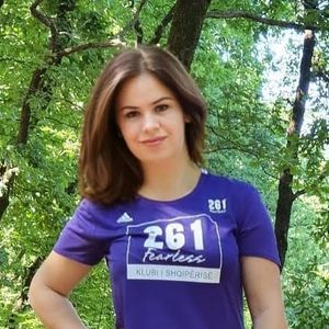A young woman in a purple "261 Fearless" T-shirt stands confidently in the shade of green trees