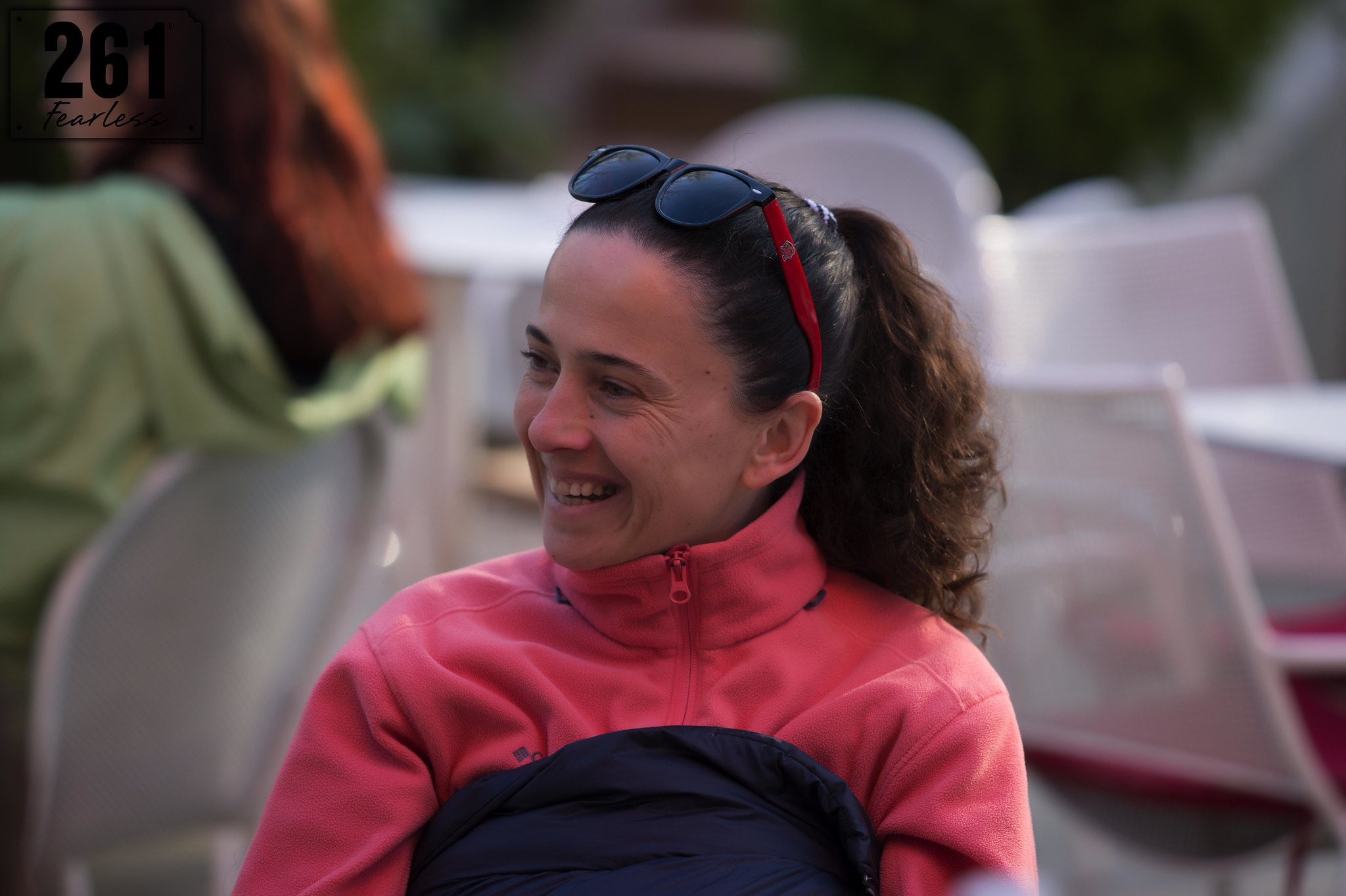 A woman from 261 Fearless Club Albania smiles warmly, wearing a pink fleece jacket and sunglasses on her head, showcasing joy and relaxation after a run