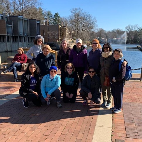 A group of ten smiling women pose together in front of a pond with a fountain and university buildings in the background, an image that represents the diversity and sense of community among the 261 Fearless running club members