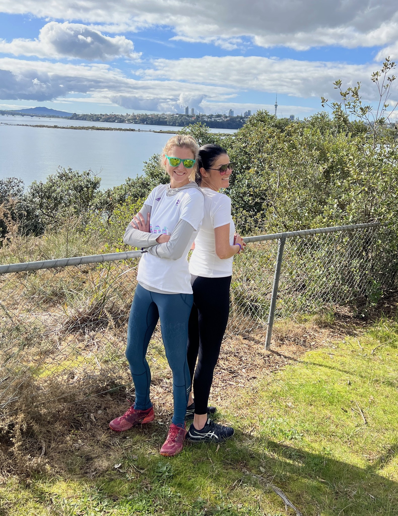 Two 261 Fearless women in sportswear lean back to back against a railing, smiling, with an idyllic view of the water and the skyline in the background 