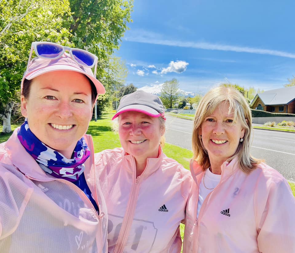 Three beaming 261 Fearless women in matching pink Adidas running jackets take a selfie, with a sunny, green landscape background