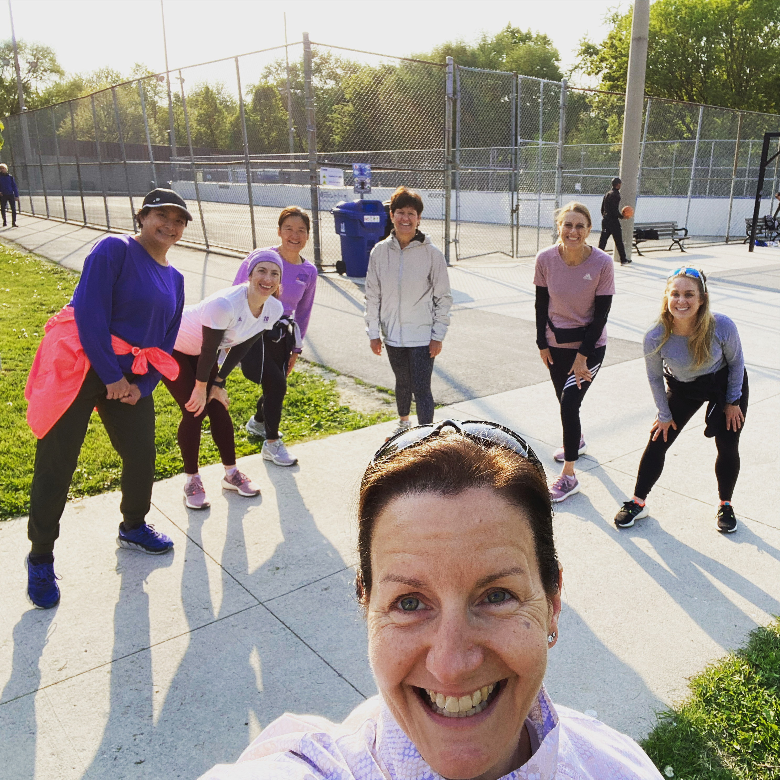 Selfie of a woman in the foreground with a group of six smiling female 261 Fearless runners in various athletic poses, ready for a run in a sunny park, exuding energy and enthusiasm