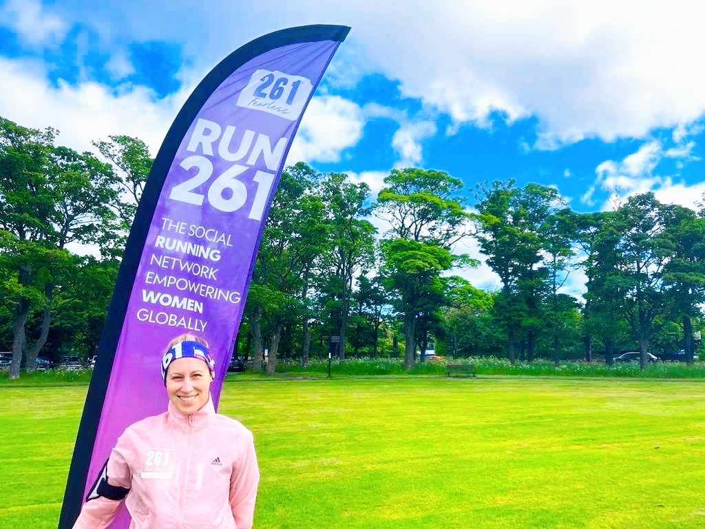Woman in pink sports jacket stands next to a 261 Fearless banner on a green field, representing the global network for women