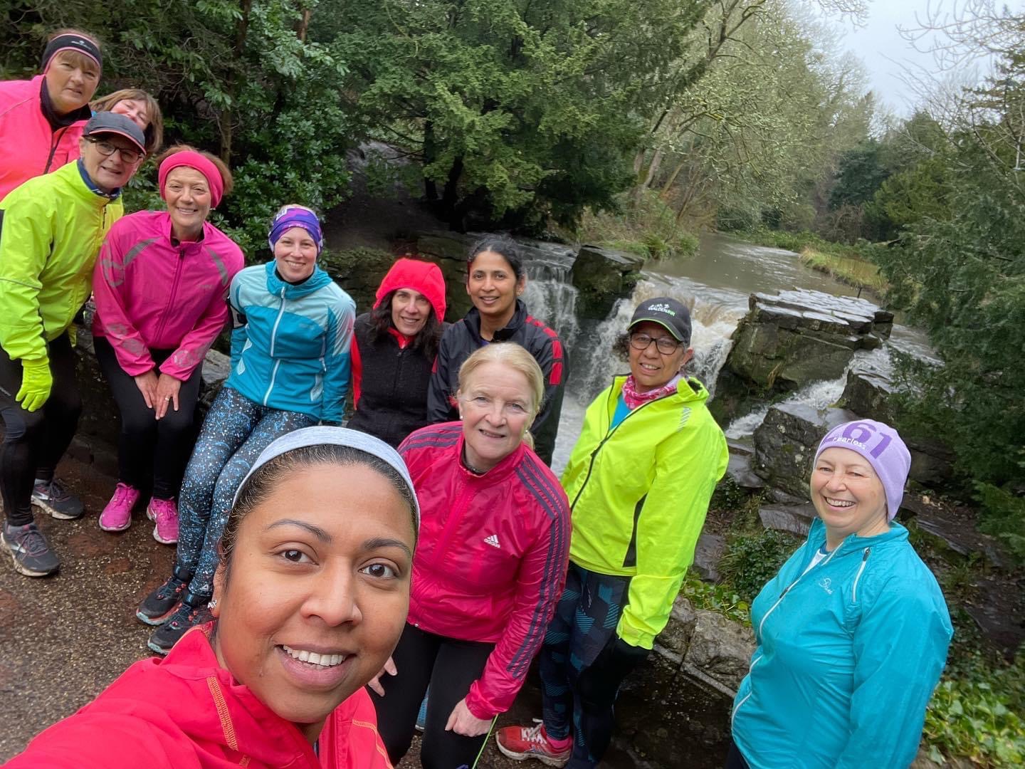 Selfie of a 261 Fearless women's running group in front of a picturesque waterfall, wearing colourful rain jackets, showing their solidarity and adventurous spirit