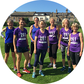 Seven women from the 261 Fearless women's running group in purple and blue running shirts stand on a green lawn, smiling and ready for a running event, with traditional buildings in the background