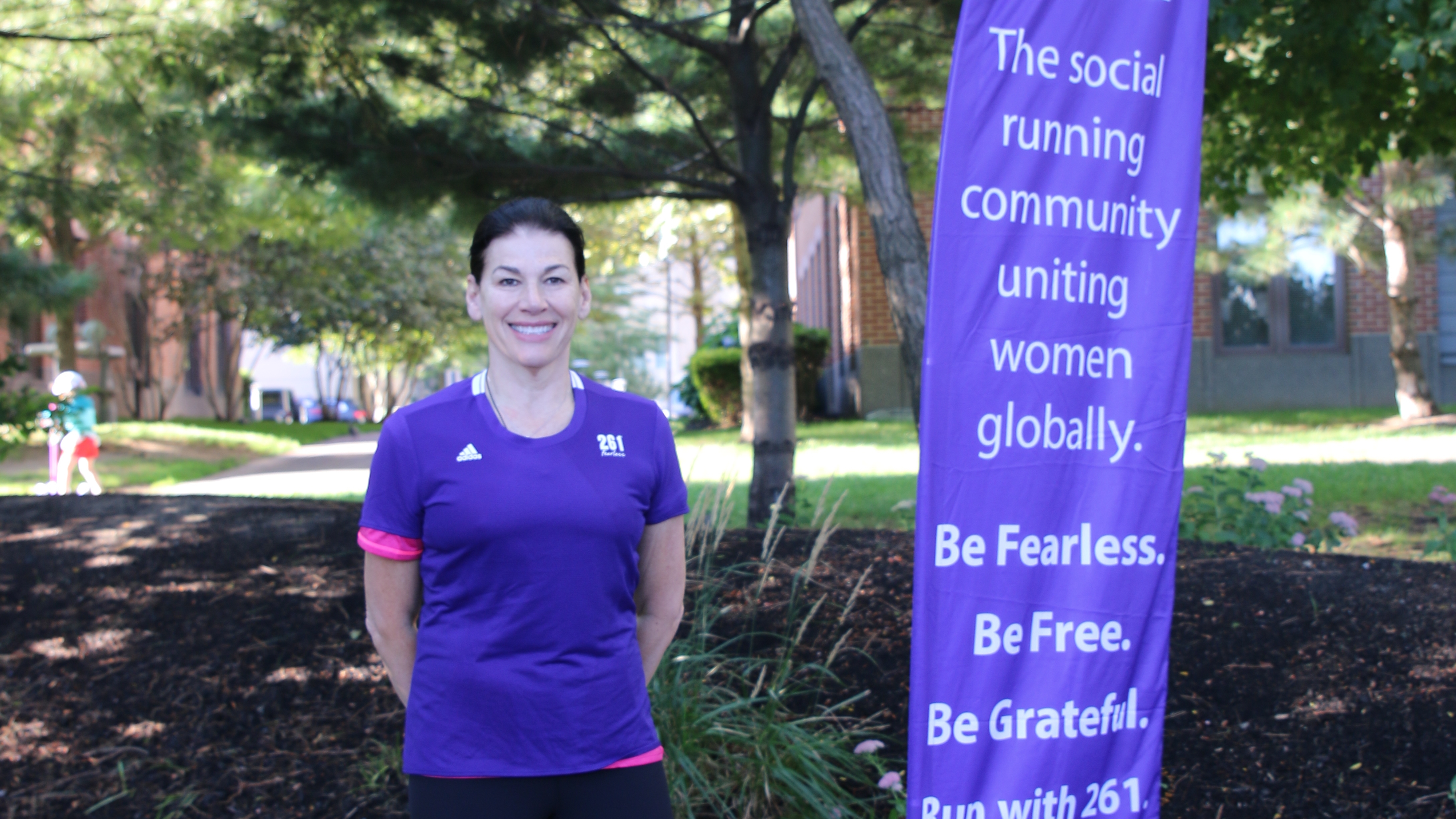 Eine Frau in einem violetten 261 Fearless Laufshirt steht lächelnd neben einem Banner der 261 Fearless Laufgruppe, das die Werte Gemeinschaft, Freiheit und Dankbarkeit betont