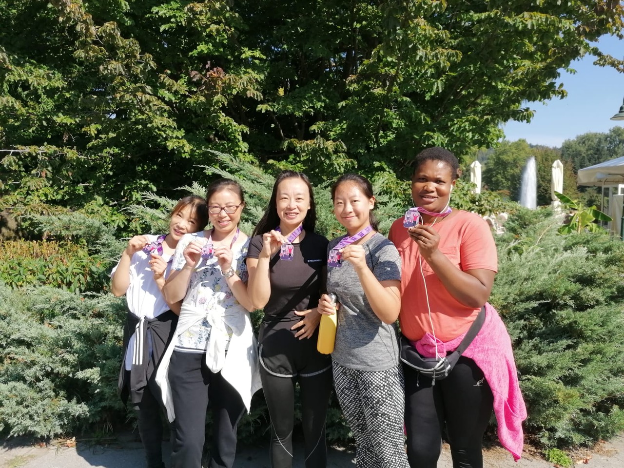 Group of women of different ethnicities, members of the 261 Fearless running group, proudly display their medals in a park-like area